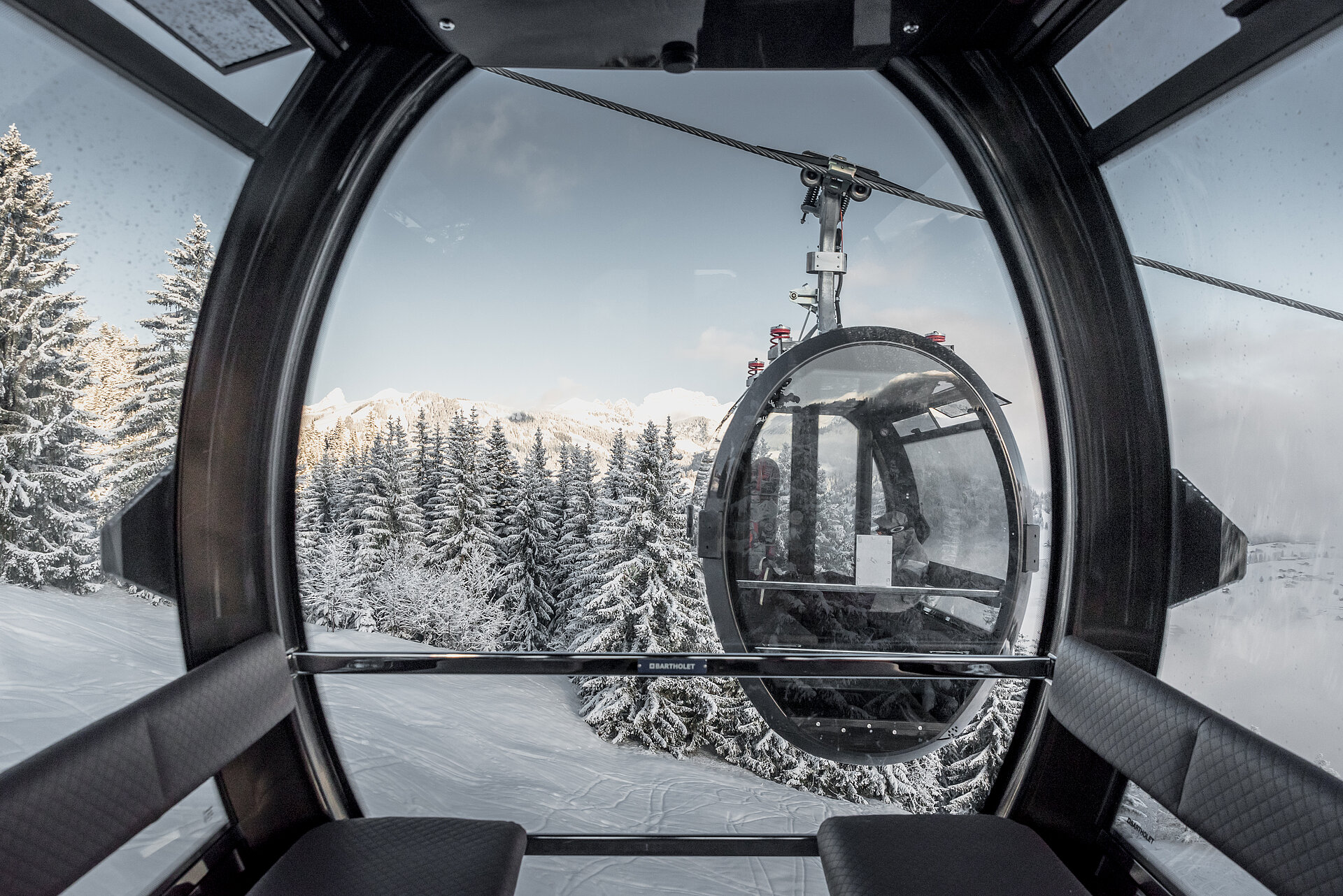 Das Foto stammt aus einer modernen 10er-Gondelbahn. Die Gondelbahn hat schwarze Lederbänke und viel Glas. Das Design ist oval und wirkt futuristisch. Ausserhalb der Gondel kreuzt eine andere Gondel mit zwei Personen darin. Draussen ist die Landschaft verschneit und nur auf den Bergspitzen sieht man die Sonne.