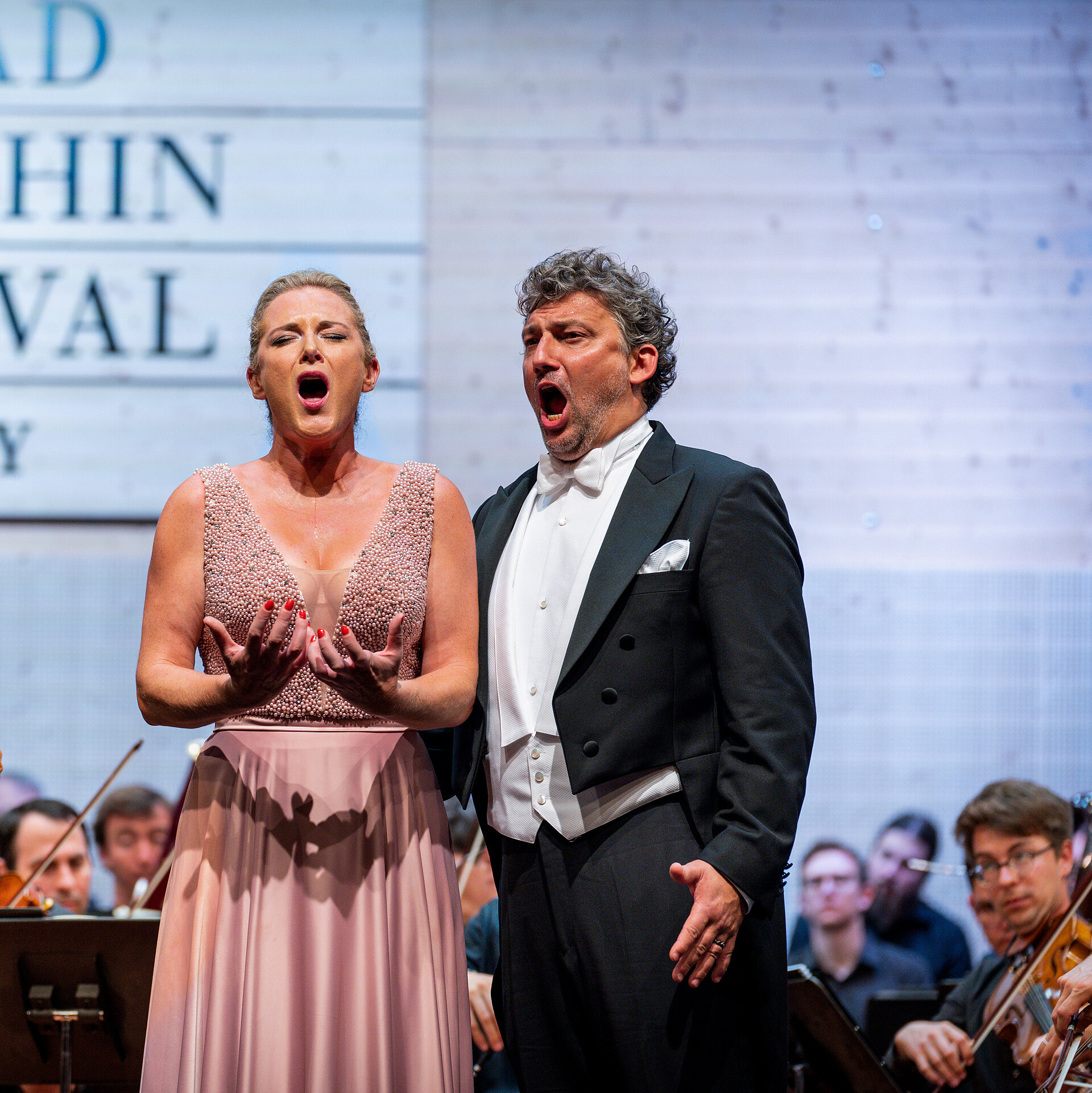 Opernsänger singen neben Dirigent voller Leidenschaft, dahinter spielt das Orchester.