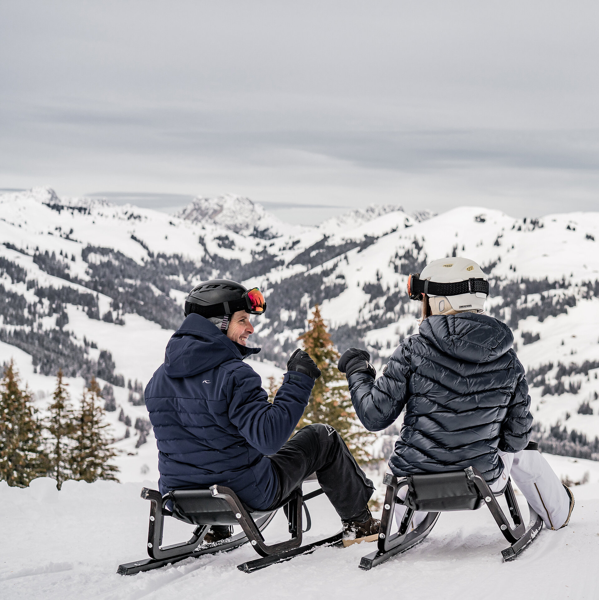 Schlittelnde Menschen in dunklen Skikleidern inmitten verschneitem Bergpanorama.