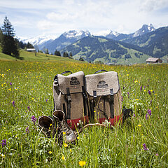 Ein Rucksack und alte Wanderschuhe, welche in hohem Grass stehen vor einer schönen Berglanschaft.